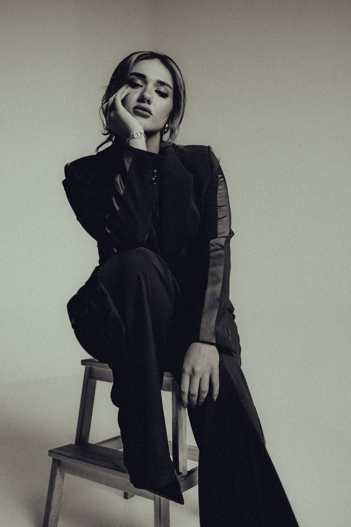 Black-and-white fashion editorial of a woman in a structured two-tone jacket seated on a wooden stool, hand resting on her cheek