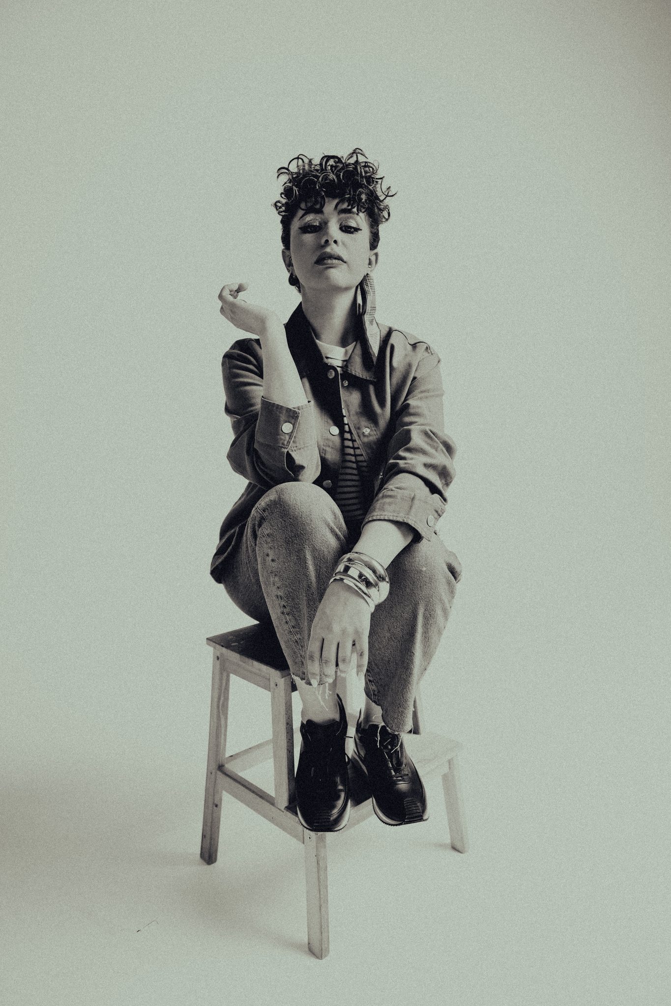 Black-and-white fashion editorial of a woman with curly hair seated cross-legged on a stool in a denim ensemble