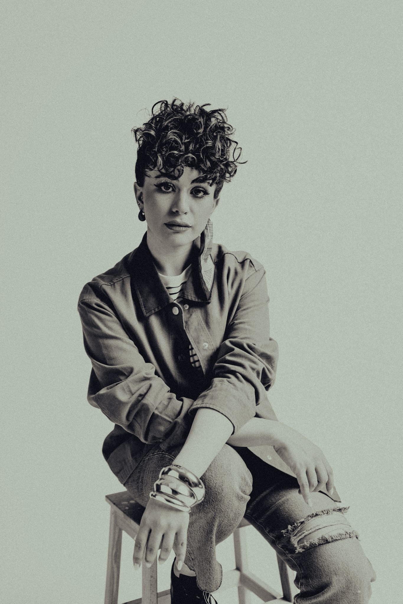 Portrait of a woman with curly hair in a denim jacket and layered bracelets seated on a stool